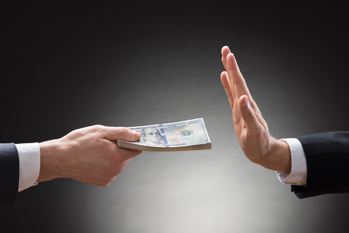 A hand offering a stack of one hundred dollar bills to a hand that makes a blocking gesture