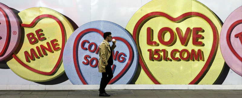A person drinks coffee as they walk in front of a wall-size advertisement.