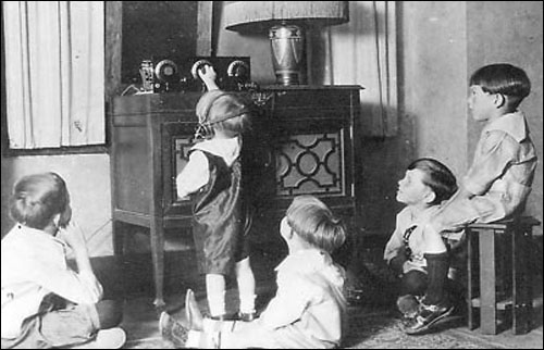 Five children are sitting and standing around a radio and listening to it