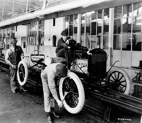 Two people are adjusting the tires on a Model T Ford on an assembly line.