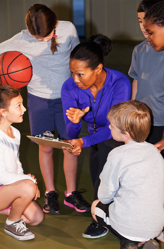 A basketball coach is kneeling, holding a clipboard, and giving directions to the young players who are huddled around her.