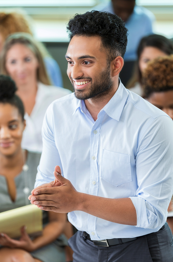 A person is speaking to a group, making eye contact and smiling, with his hands pressed together.