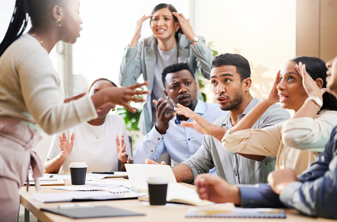 A group of colleagues are arguing. They are sitting around a table, gesturing with their hands and looking upset