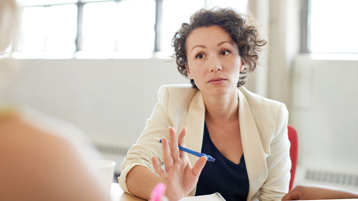A person with a notebook and pen is mediating a discussion between two other people. She looks serious and her hand is slightly raised.